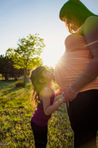 A small child gently leands against the belly of a pregnant woman.